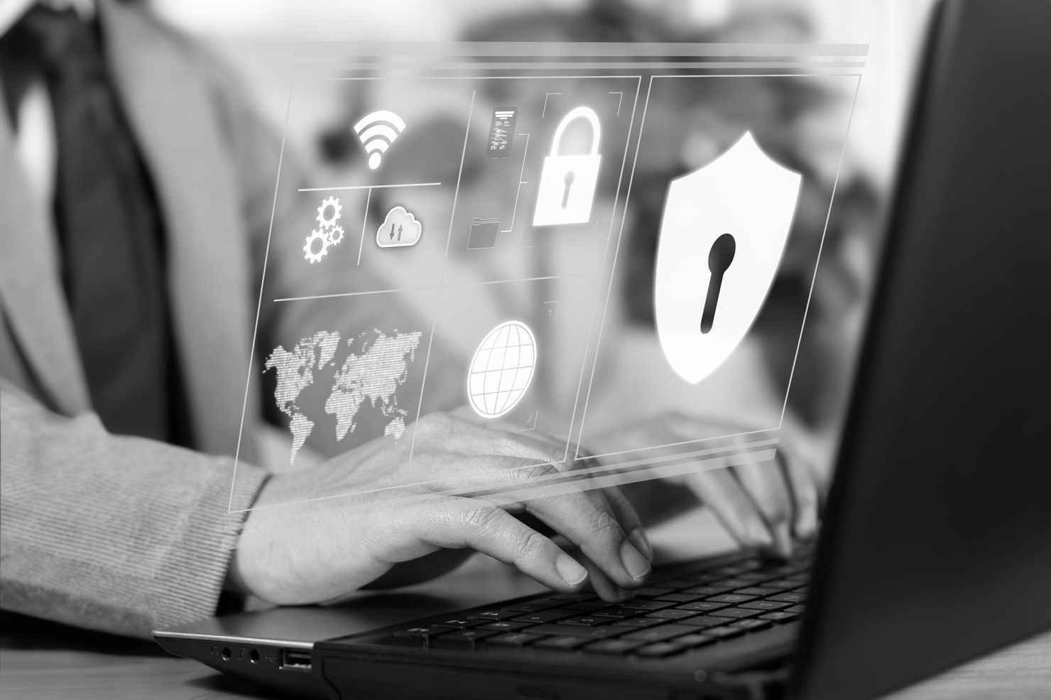 Person typing on laptop with holographic icons of a lock, shield, globe, and gear in grayscale, suggesting cybersecurity themes.
