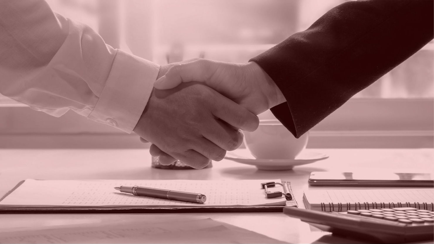 Two people shaking hands over a desk with papers, pen, notebook, and calculator. Sunlit background with a cup in sepia tone.
